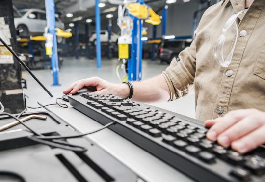 Caucasian Automotive Technician Checking Vehicle Database Documentation. Hands on Computer Keyboard Inside Auto Service Station.