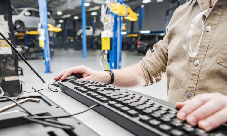 Caucasian Automotive Technician Checking Vehicle Database Documentation. Hands on Computer Keyboard Inside Auto Service Station.