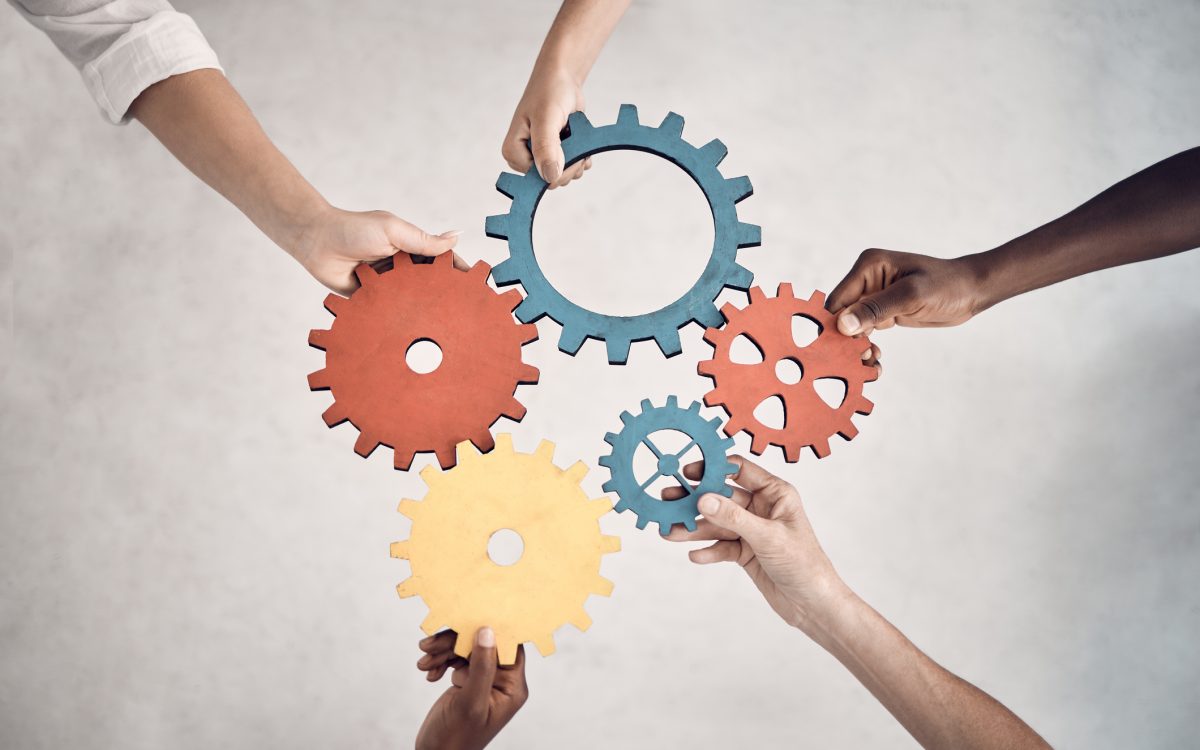 Group of five businesspeople holding and fitting gears together in an office at work. United business professionals having fun connecting gear pieces during a meeting