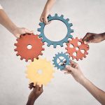Group of five businesspeople holding and fitting gears together in an office at work. United business professionals having fun connecting gear pieces during a meeting
