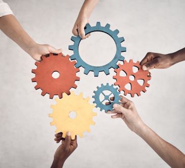 Group of five businesspeople holding and fitting gears together in an office at work. United business professionals having fun connecting gear pieces during a meeting
