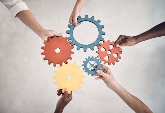 Group of five businesspeople holding and fitting gears together in an office at work. United business professionals having fun connecting gear pieces during a meeting