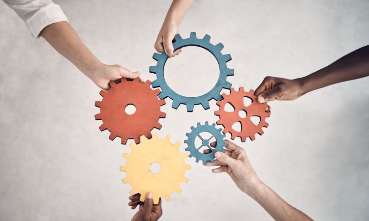 Group of five businesspeople holding and fitting gears together in an office at work. United business professionals having fun connecting gear pieces during a meeting