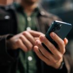 Close-up hands of unrecognizable man holding and using smartphone standing on city street, browsing internet, checking social media, using mobile application. Concept of modern communication.