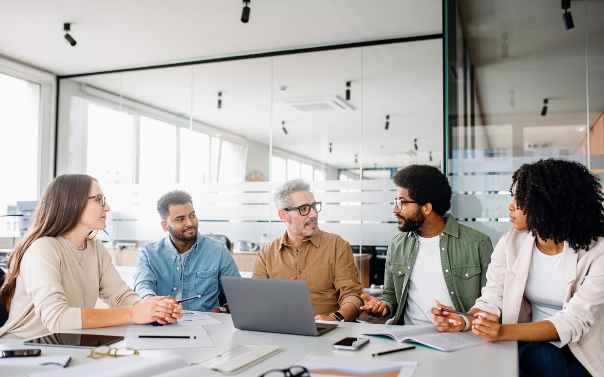 A diverse office team is engaged in a strategic meeting, with a senior colleague leading discussion, illustrating a productive and inclusive workplace. Colleagues engage in a lively exchange of ideas