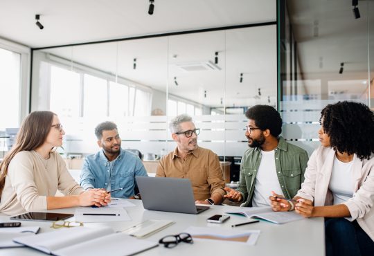 A diverse office team is engaged in a strategic meeting, with a senior colleague leading discussion, illustrating a productive and inclusive workplace. Colleagues engage in a lively exchange of ideas