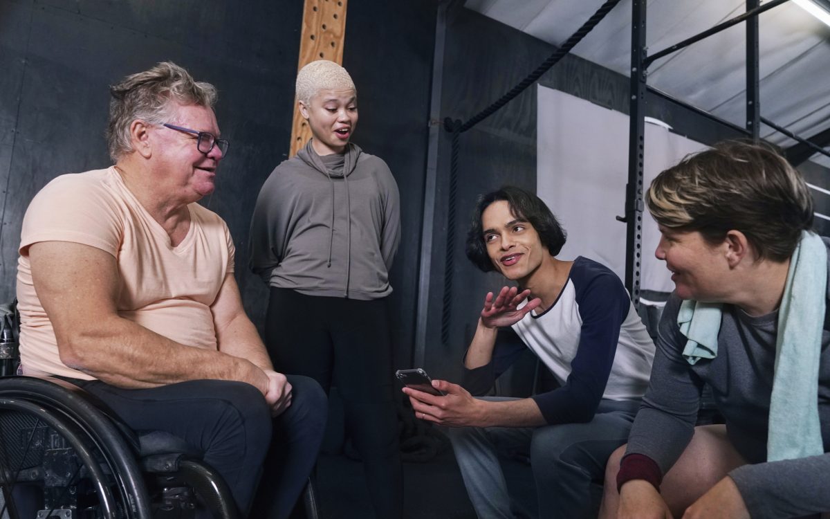 Smiling friends, including man in wheelchair, sitting and talking in gym after workout