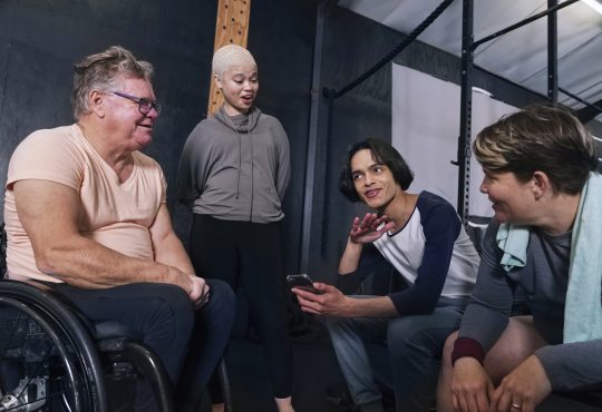 Smiling friends, including man in wheelchair, sitting and talking in gym after workout
