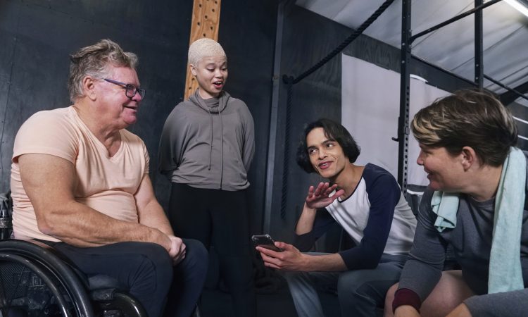 Smiling friends, including man in wheelchair, sitting and talking in gym after workout