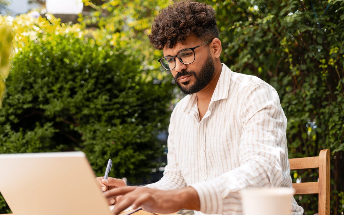 Young businessman taking notes while working remotely on a laptop at a garden cafe, enjoying the vibrant outdoor atmosphere
