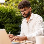 Young businessman taking notes while working remotely on a laptop at a garden cafe, enjoying the vibrant outdoor atmosphere