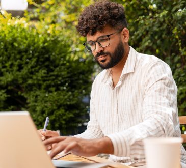 Young businessman taking notes while working remotely on a laptop at a garden cafe, enjoying the vibrant outdoor atmosphere