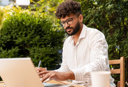Young businessman taking notes while working remotely on a laptop at a garden cafe, enjoying the vibrant outdoor atmosphere