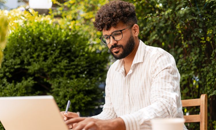 Young businessman taking notes while working remotely on a laptop at a garden cafe, enjoying the vibrant outdoor atmosphere