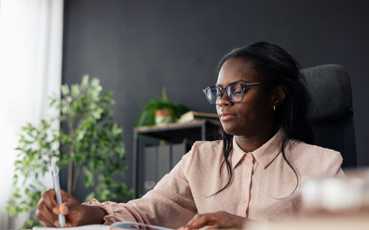 Black woman student wearing glasses writing notes in a notebook at an office desk
