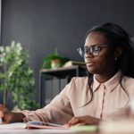 Black woman student wearing glasses writing notes in a notebook at an office desk