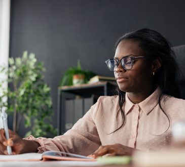 Black woman student wearing glasses writing notes in a notebook at an office desk