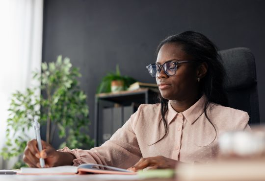 Black woman student wearing glasses writing notes in a notebook at an office desk