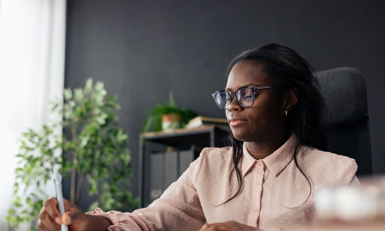 Black woman student wearing glasses writing notes in a notebook at an office desk