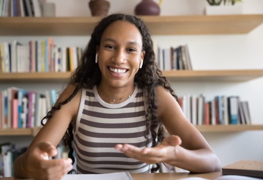 Webcam view of African student girl wear earphones looking at camera and speaking, participate in education webinar, talking with tutor, discussing joint schoolwork with groupmate using video call app