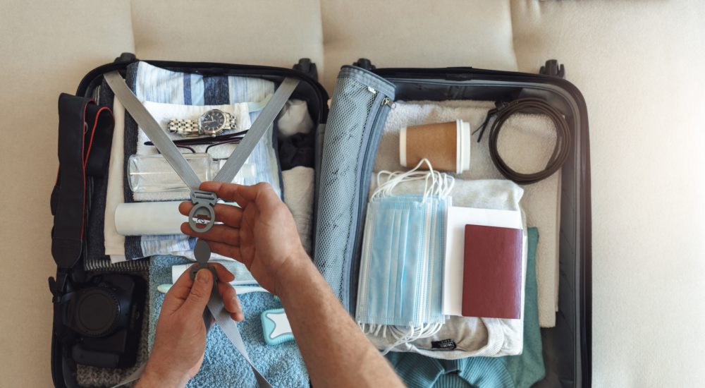 Men's hands putting a face mask and passport in the suitcase prepared to travel, in the new normal, after the coronavirus COVID-19 pandemic. High angle view of suitcase with clothes, passport and masks