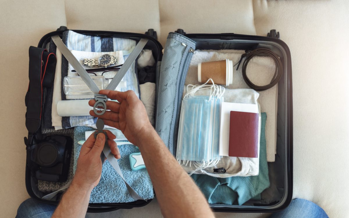 Men's hands putting a face mask and passport in the suitcase prepared to travel, in the new normal, after the coronavirus COVID-19 pandemic. High angle view of suitcase with clothes, passport and masks
