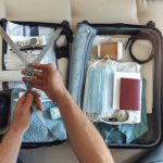 Men's hands putting a face mask and passport in the suitcase prepared to travel, in the new normal, after the coronavirus COVID-19 pandemic. High angle view of suitcase with clothes, passport and masks