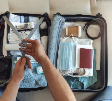 Men's hands putting a face mask and passport in the suitcase prepared to travel, in the new normal, after the coronavirus COVID-19 pandemic. High angle view of suitcase with clothes, passport and masks