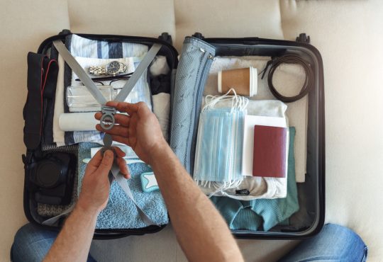 Men's hands putting a face mask and passport in the suitcase prepared to travel, in the new normal, after the coronavirus COVID-19 pandemic. High angle view of suitcase with clothes, passport and masks