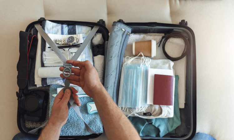 Men's hands putting a face mask and passport in the suitcase prepared to travel, in the new normal, after the coronavirus COVID-19 pandemic. High angle view of suitcase with clothes, passport and masks
