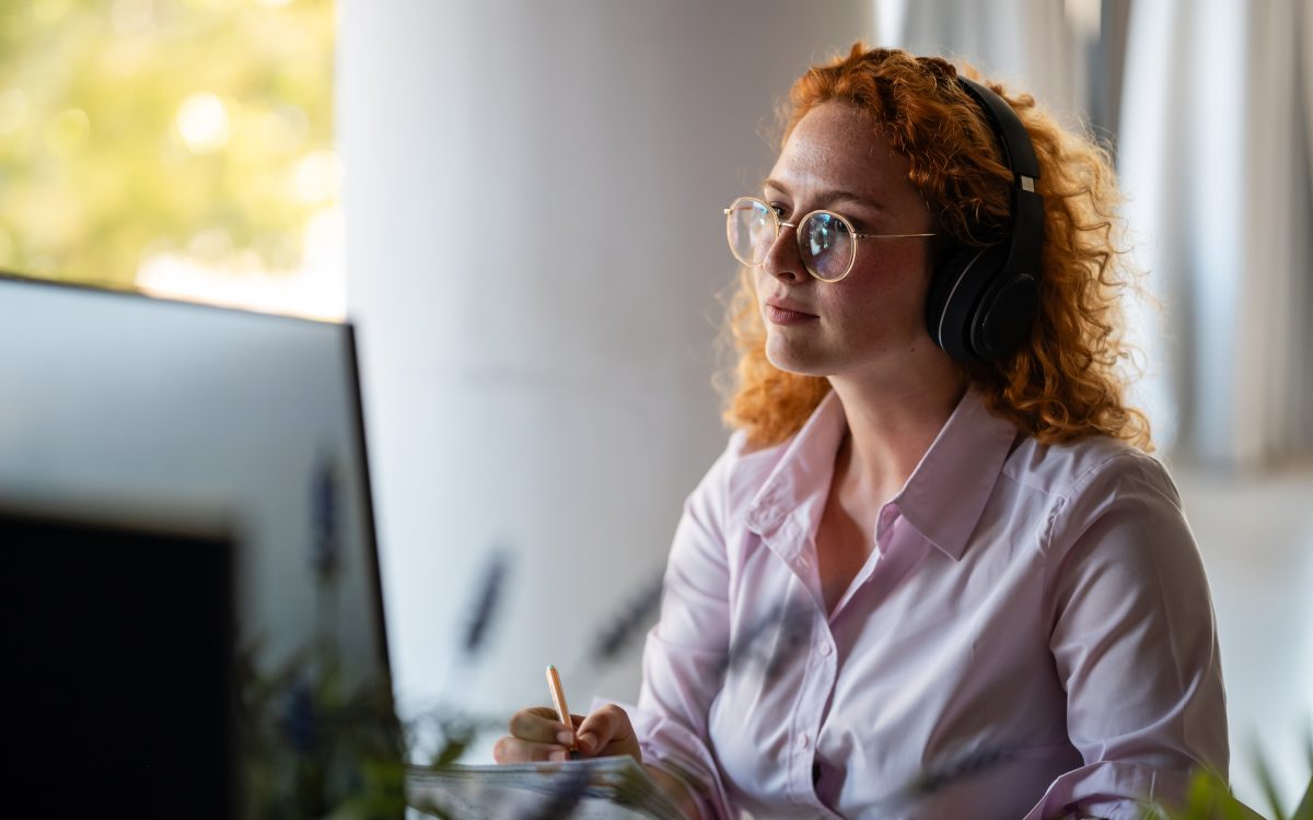 Concentrated young woman with curly red hair wearing headphones and glasses, working at computer in bright modern workspace