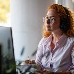 Concentrated young woman with curly red hair wearing headphones and glasses, working at computer in bright modern workspace