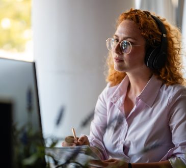 Concentrated young woman with curly red hair wearing headphones and glasses, working at computer in bright modern workspace