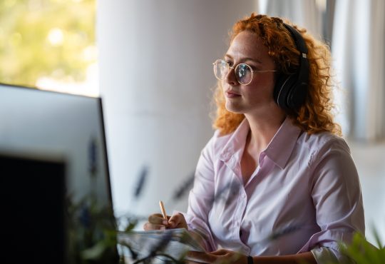Concentrated young woman with curly red hair wearing headphones and glasses, working at computer in bright modern workspace