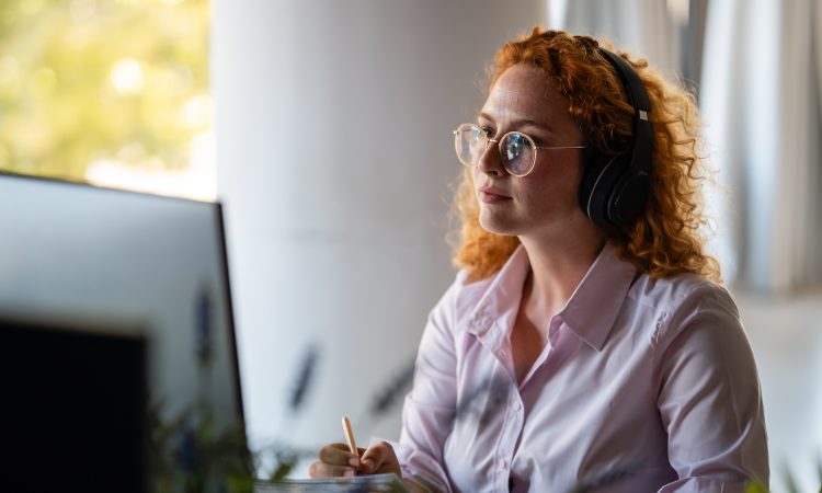 Concentrated young woman with curly red hair wearing headphones and glasses, working at computer in bright modern workspace