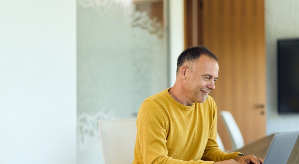 Mature man in a yellow sweater types on a laptop at a modern home office table, smiling while focused on a virtual meeting; ideal for remote work, productivity, and corporate communication content.
