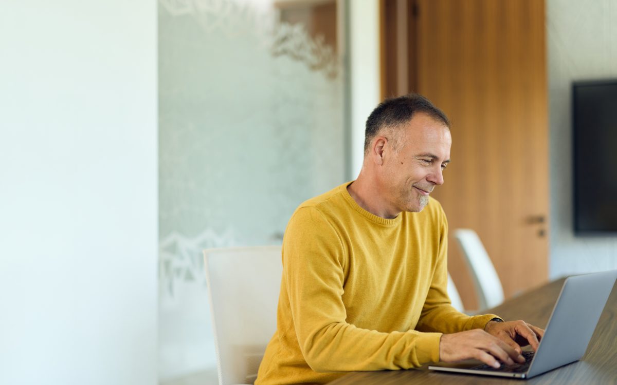 Mature man in a yellow sweater types on a laptop at a modern home office table, smiling while focused on a virtual meeting; ideal for remote work, productivity, and corporate communication content.