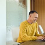Mature man in a yellow sweater types on a laptop at a modern home office table, smiling while focused on a virtual meeting; ideal for remote work, productivity, and corporate communication content.