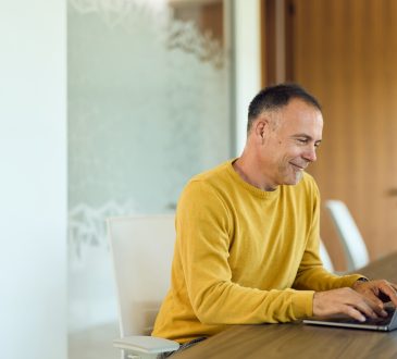 Mature man in a yellow sweater types on a laptop at a modern home office table, smiling while focused on a virtual meeting; ideal for remote work, productivity, and corporate communication content.