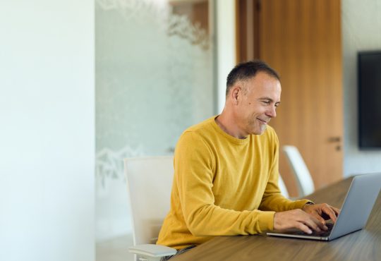 Mature man in a yellow sweater types on a laptop at a modern home office table, smiling while focused on a virtual meeting; ideal for remote work, productivity, and corporate communication content.