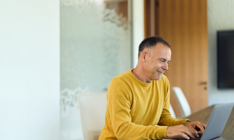 Mature man in a yellow sweater types on a laptop at a modern home office table, smiling while focused on a virtual meeting; ideal for remote work, productivity, and corporate communication content.