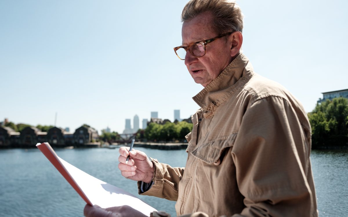 Senior engineer examining architectural plans near the river, with the iconic canary wharf skyline in the background, enjoying a sunny day in vibrant london