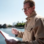 Senior engineer examining architectural plans near the river, with the iconic canary wharf skyline in the background, enjoying a sunny day in vibrant london