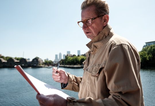 Senior engineer examining architectural plans near the river, with the iconic canary wharf skyline in the background, enjoying a sunny day in vibrant london
