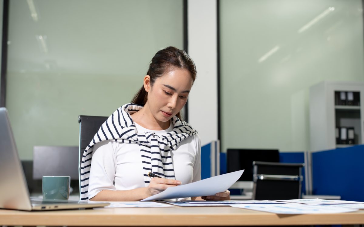 Young woman working at an office desk, reading and reviewing papers with a laptop nearby in a modern workspace