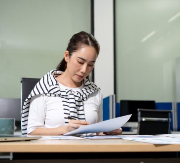 Young woman working at an office desk, reading and reviewing papers with a laptop nearby in a modern workspace