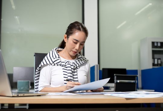 Young woman working at an office desk, reading and reviewing papers with a laptop nearby in a modern workspace
