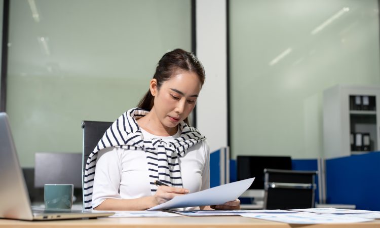 Young woman working at an office desk, reading and reviewing papers with a laptop nearby in a modern workspace
