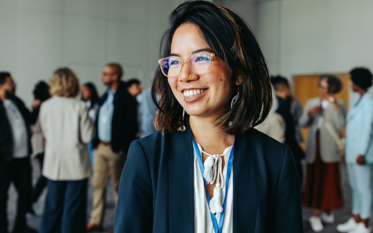 Asian businesswoman in glasses, smiling and networking at a professional conference. Colleagues in the background enhance the atmosphere of collaboration and connection.