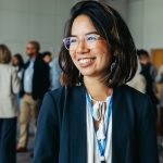 Asian businesswoman in glasses, smiling and networking at a professional conference. Colleagues in the background enhance the atmosphere of collaboration and connection.